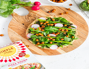 Spring vegetable pizza on round wooden plate on countertop with Simple Mills pizza dough box