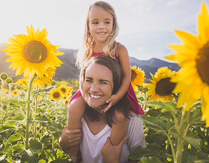 Daughter sitting on mothers shoulders walking in sunflower patch