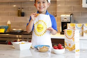 Young smiling boy holding up a Simple Mills Farmhouse Cheddar Almond Flour Cracker in the kitchen